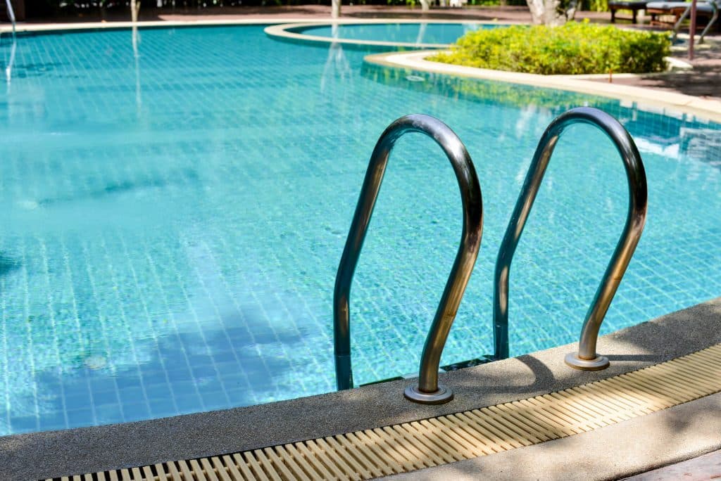 Curved stainless steel ladder rails at the edge of a blue tiled pool, next to a slotted gutter grate and gray concrete deck
