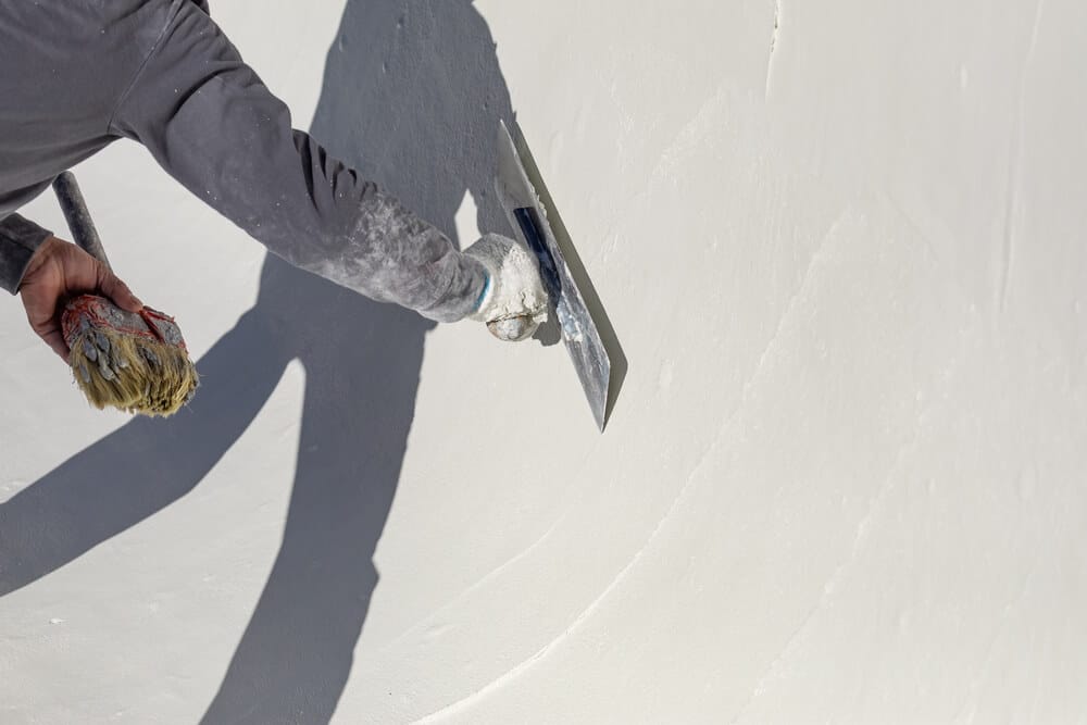 Worker in gray sleeves smoothing fresh white plaster on a pool wall with a steel trowel, holding a brush in the other hand