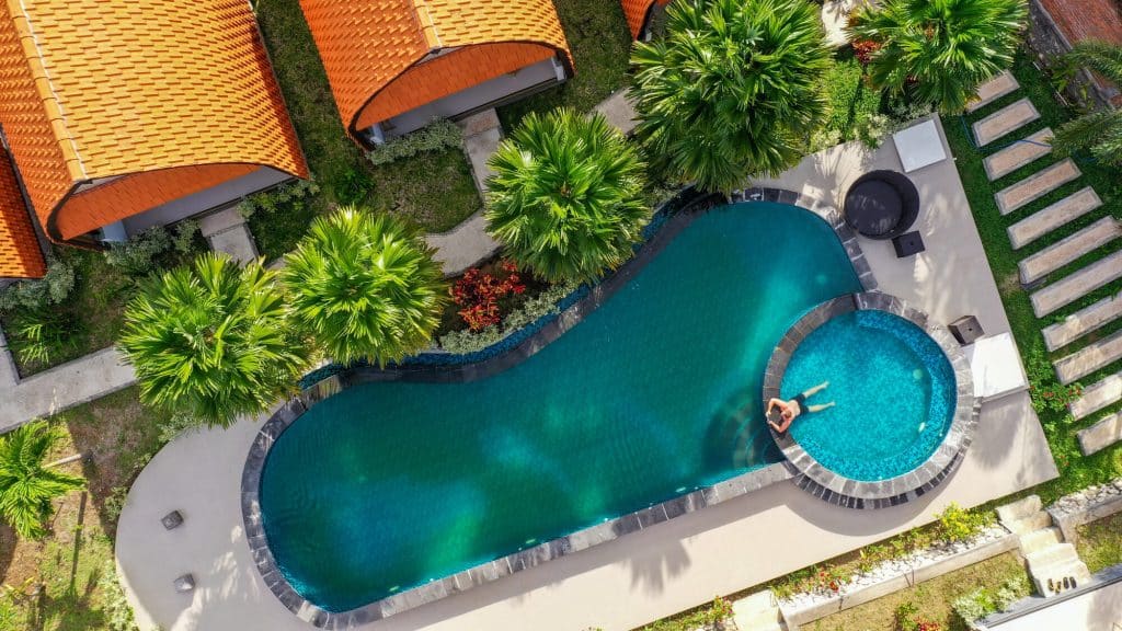 Aerial view of a curving free-form pool with an attached round spa, dark stone coping, and palm trees around a light concrete deck