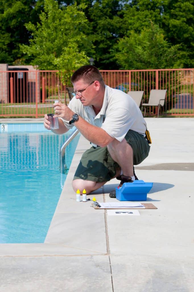 Man kneeling at the edge of a pool holding up a water sample vial, with a blue test kit, reagent bottles, and a clipboard on the deck beside him