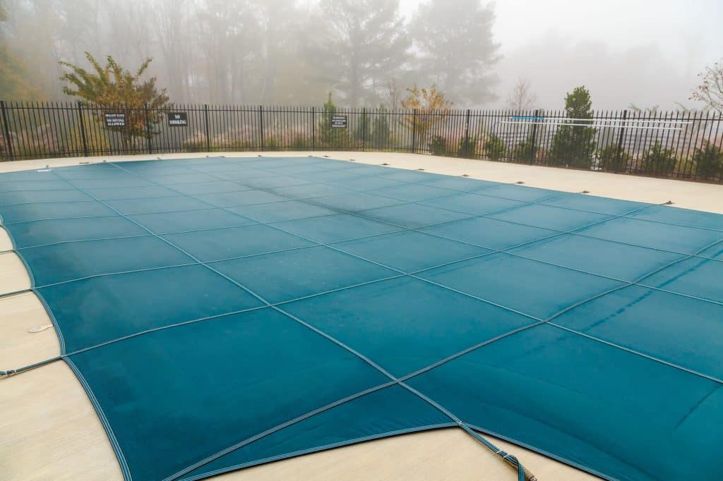 Teal mesh safety cover stretched tight across an inground pool on a foggy morning, with a black metal fence behind it