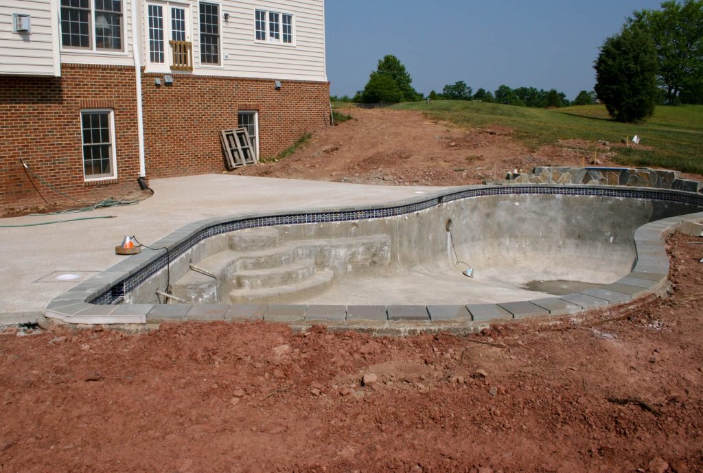 Unfinished kidney-shaped concrete pool shell with bare walls and built-in steps, a blue waterline tile band, and gray coping, surrounded by bare dirt beside a brick house