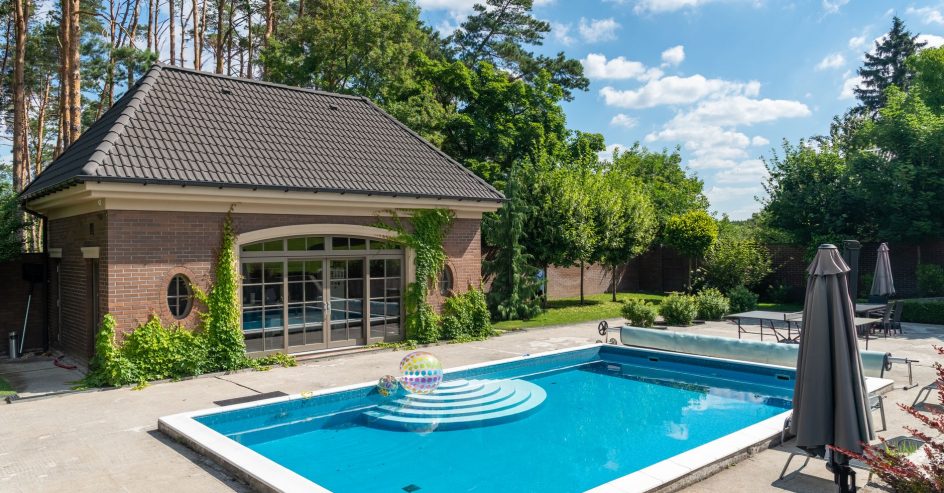 Finished rectangular backyard pool with curved entry steps beside a brick pool house under tall pines
